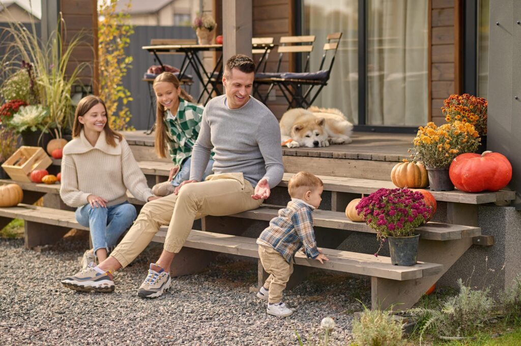 Eine Familie sitzt auf der Veranda und beobachtet ein kleines Kind, das auf Holzstufen krabbelt. Im Hintergrund sind Herbstdekorationen und ein schlafender Hund zu sehen. Eine Schließanlage sorgt dafür, dass die verschiedenen Bereiche des Hauses sicher bleiben.