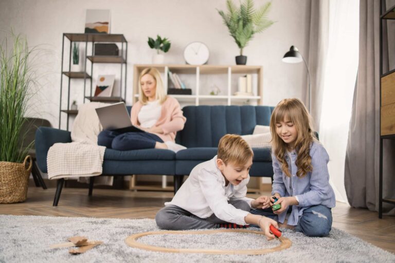 Zwei Kinder spielen auf einem Teppich im Wohnzimmer, während eine Frau im Hintergrund auf dem Sofa am Laptop arbeitet – sicheres Zuhause dank moderner Schließanlage im Privathaushalt.
